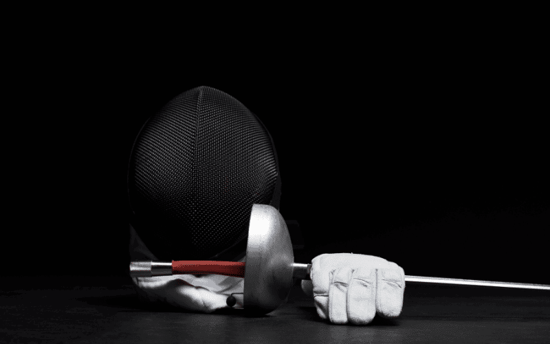 A set of beginner's fencing gear laid out neatly on a wooden table, including a mask, jacket, glove, and foil